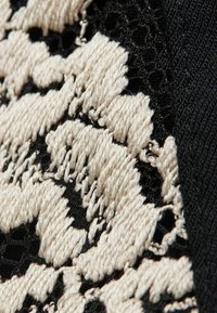 Texturerad svart tyg med ett krämfärgat, upphöjt abstrakt mönster. Designen inkluderar intrikata broderier med en spetsbakgrund.