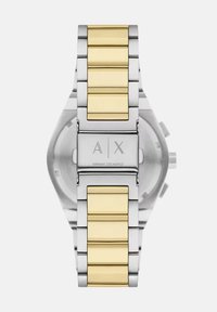 Reloj de pulsera de metal plateado y dorado con una correa de acero inoxidable y acentos en amarillo. Presenta un diseño minimalista y un logotipo grabado en el cierre.