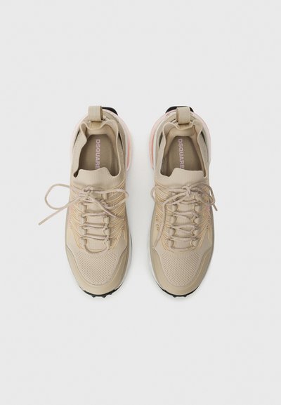 Scarpe atletiche beige con una tomaia in mesh testurizzato, lacci piatti e una suola arrotondata. Presentano un accento rosa e nero sul tallone.