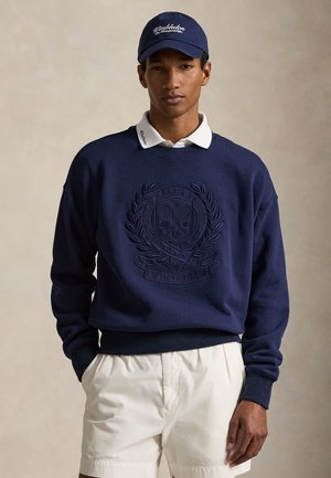 Homme portant une casquette de baseball bleu marine Wimbledon, un sweat-shirt bleu marine avec l'écusson Ralph Lauren Wimbledon, une chemise blanche à col et un short blanc.