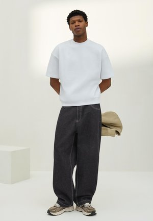 Mladý muž stojí s rukami za chrbtom, má na sebe jednoduchú bielu nadmernú krátku košeľu, voľné tmavé džínsy, béžové tenisky a drží béžovú bundu.