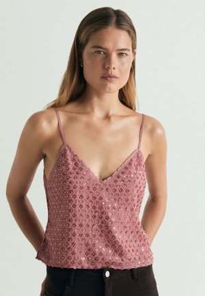 Mujer con cabello largo y castaño lleva una blusa rosa sin mangas decorada con pequeñas cuentas y lentejuelas, combinada con pantalones oscuros, de pie sobre un fondo blanco.
