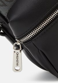 Borsa in pelle nera con dettagli in argento sulla cerniera e un'etichetta di branding. Texture liscia con una forma strutturata e arrotondata.