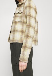 Brun- och gulrutigt jacka, kort design, mjuk textur, svarta knappar, två framfickor och krage. Bärs med olivfärgade byxor.