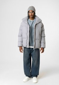 Grijze pufferjack met een hoge kraag, dikke vulling en ritssluiting, gedragen over een lichtblauw sweatshirt en donkere wijde jeans.