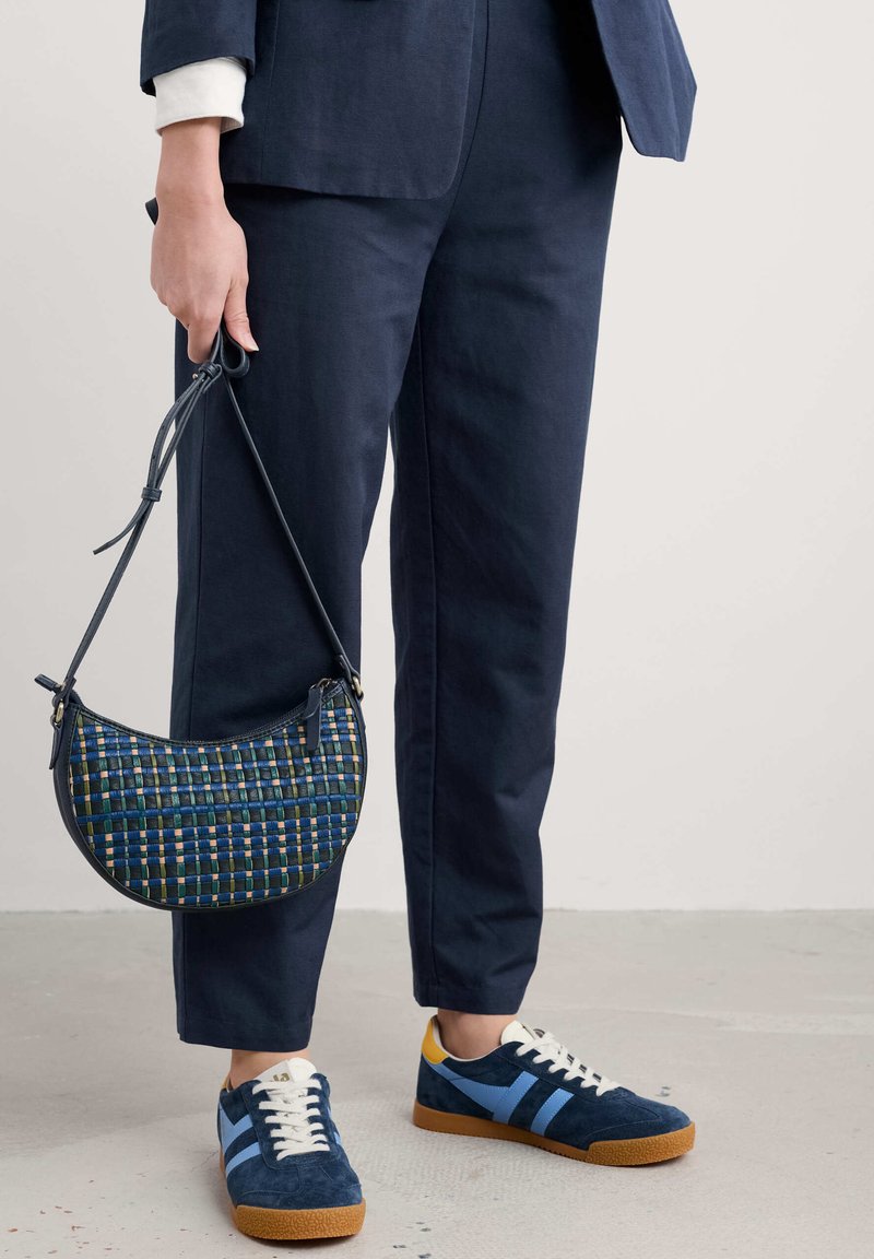 Bolso en forma de media luna con un patrón tejido en azul, verde y amarillo. Combinado con pantalones marino y zapatillas de ante azules con acentos blancos.