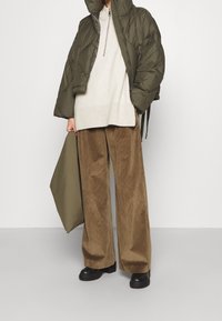 Zelená péřová bunda, krémový svetr a hnědé široké manšestrové kalhoty. Černé platformové boty. Drží olivově zelenou kabelku.