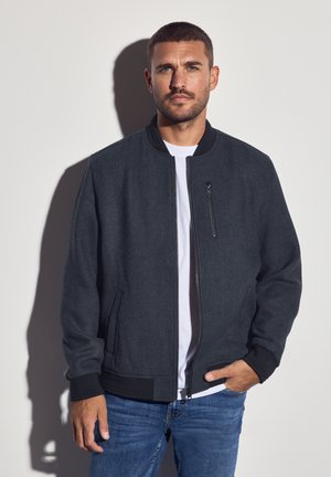 Uomo con capelli corti e barba che indossa una giacca grigia scura con zip, una maglietta bianca e jeans blu, in piedi contro un muro chiaro e uniforme.