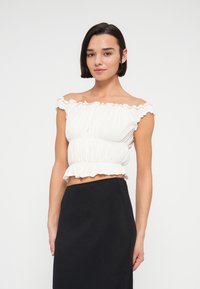 Haut court blanc à épaules dénudées avec bordure à volants, texture froncée et taille ajustée, associé à une jupe noire. Finition satinée sur le haut.