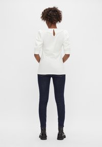 Top blanco con mangas abullonadas y cuello con estrías, con un escote en forma de ojo de cerradura en la parte de atrás, combinado con jeans ajustados oscuros y botas negras.