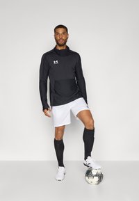 Black long-sleeve sports top with a high collar, paired with white shorts, black compression sleeves, and white soccer shoes. Black and white soccer ball.