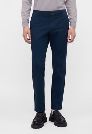 Mann trägt dunkelblaue Slim-Fit-Hose, schwarze Loafers, weiße Socken und ein hellgraues Langarmoberteil und steht vor einem einfarbigen Hintergrund.