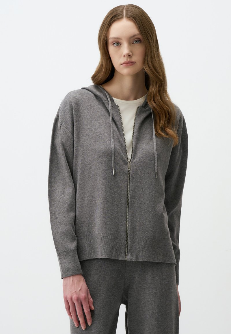 Haut à zip gris avec capuche, doté d'une texture douce, d'un ourlet côtelé et de manches longues. Associé à un pantalon assorti, offrant une coupe décontractée.