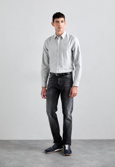 Light gray button-up shirt, dark gray jeans, and navy sneakers with white laces. Man stands on concrete floor against a gray wall.