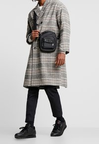 Black crossbody bag with a smooth texture, featuring a front pocket and green accents, worn over a gray plaid coat and dark tapered pants.