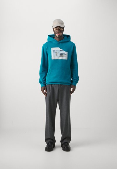 Teal hoodie with a front pocket and graphic print. Paired with gray, loose-fitting trousers and black shoes. Cap in light beige.
