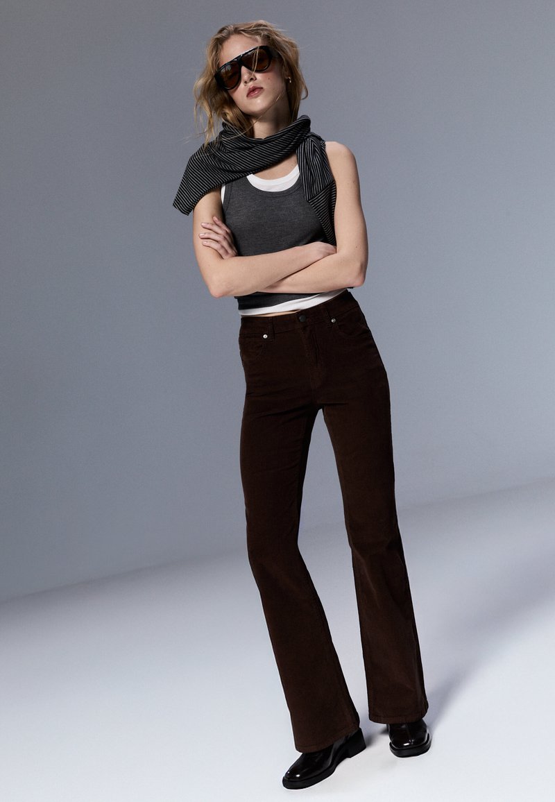 Pantalon évasé en velours côtelé marron avec poches avant, associé à un débardeur gris et une écharpe rayée noir et blanc drapée autour du cou.