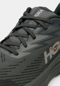 Κοντινή λήψη ενός γκρι παπουτσιού τρεξίματος Hoka με μαύρα κορδόνια και υφασμάτινη πλέξη με υφή, παρουσιάζοντας το λογότυπο της μάρκας στην πλευρά.