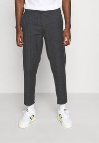 Pantalon à carreaux gris avec une coupe ajustée, présentant une texture subtile et des jambes fuselées, associé à des baskets blanches avec des accents de couleur.