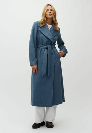 Woman with blonde hair wears long blue belted coat, white pants, and dark shoes, standing against plain light background.