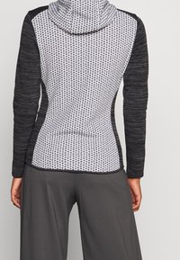 Achteraanzicht van een persoon in een getailleerde hoodie met een zwart-wit patroon op het achterpaneel en zwarte textuur mouwen, gecombineerd met donkere broeken.