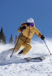 Gele skikleding met zwarte accenten, witte helm, paarse skibril en skistokken, skiën op sneeuw met bomen op de achtergrond en een heldere blauwe lucht.