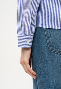 Χέρι που ακουμπά δίπλα στην τσέπη μπλε τζιν παντελονιού, φορώντας μπλε και λευκό πουκάμισο με κάθετες ρίγες και μανίκι με κουμπί.