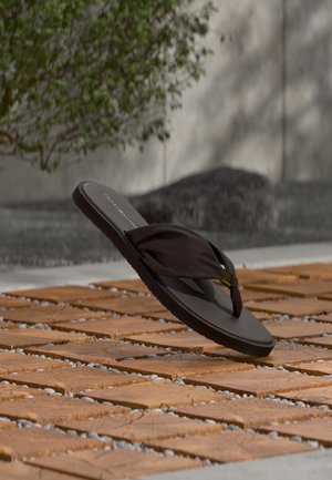 Tongs noires avec brides en tissu posées sur un sol en dalles de bois extérieur, avec des plantes floues et une pierre en arrière-plan.