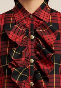 Camicia a scacchi rossa e nera con linee gialle, dotata di bottoni dorati e tessuto arricciato che scende al centro anteriore sotto il colletto.