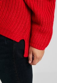 Czerwona sweter dziany z teksturowanym wzorem, ściągaczami na rękawach i rozcięciem po boku, w zestawieniu z ciemnym dżinsem. Zbliżenie na rękaw i dłoń.