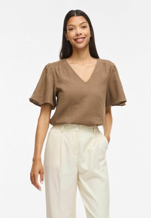 Femme souriante, portant un chemisier marron à manches courtes avec col en V et un pantalon large crème taille haute, debout devant un fond blanc uni.