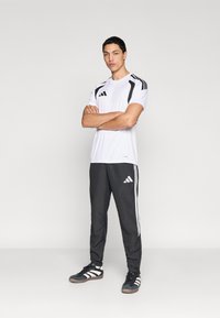 Λευκό αθλητικό πουκάμισο με μαύρες λεπτομέρειες, συνδυασμένο με μαύρο παντελόνι φόρμας με λευκές ρίγες. Το μοντέλο φοράει μαύρα αθλητικά παπούτσια.