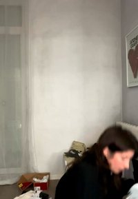 Γυναίκα με σκούρα μαλλιά κάθεται στο πάτωμα σε ένα δωμάτιο με λευκό τοίχο, καλοριφέρ, αφίσα με κόκκινη καρδιά και διασκορπισμένα ρούχα και κουτιά.