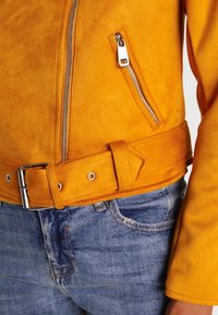 Gul mockajacka med en ficka som stängs med dragkedja, midja med bälte och dekorativa sömmar. Bärs över blå denimjeans.