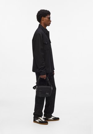 Black jacket with a button closure, patterned black pants, and black and white sneakers. A small black bag with graphic detail is held.