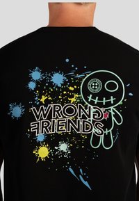 Zwarte lange mouwen shirt met een kleurrijk grafisch ontwerp met spetterpatronen. Tekst "WRONG FRIENDS" en een cartoonfiguur zijn afgebeeld.