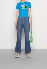 Camiseta gráfica azul de manga corta, combinada con unos vaqueros de mezclilla azules acampanados. Sosteniendo un pequeño bolso verde y usando zapatillas deportivas de color claro.
