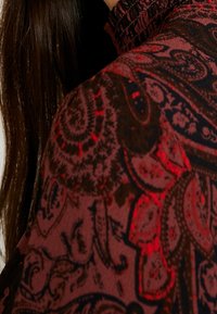 Una tela de color rojo oscuro con un patrón presenta intrincados diseños de paisley. La textura parece suave, con un cuello rizado que añade detalle.