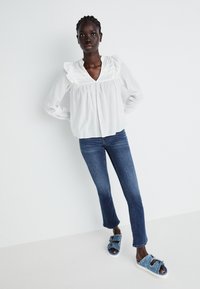 White long-sleeve blouse with ruffled neckline, paired with fitted blue jeans and blue slides featuring a decorative strap detail.