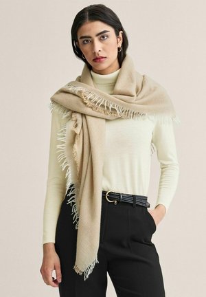 Kvinde iført beige frynset tørklæde over cremefarvet rullekravesweater, sorte bukser med vævet bælte, stående med hånden i lommen mod en ensfarvet baggrund.