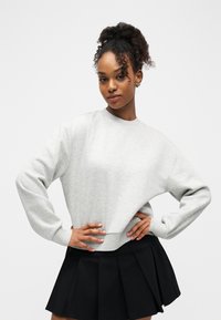Jeune femme aux cheveux bouclés coiffés en chignon haut, portant un sweat-shirt gris clair et une jupe plissée noire, posant les mains sur les hanches.