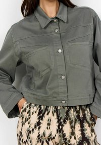 Femme portant une veste verte courte à boutons avec des poches avant et un pantalon beige taille haute avec des motifs abstraits vert foncé et noir.