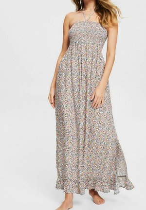 Femme portant une robe maxi fleurie sans manches à encolure américaine avec un corsage smocké et un ourlet à volants, debout pieds nus sur un fond uni.