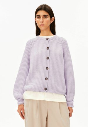 Femme portant un cardigan en maille lilas boutonné sur une chemise blanche avec un pantalon beige à jambes larges, debout devant un fond blanc uni.