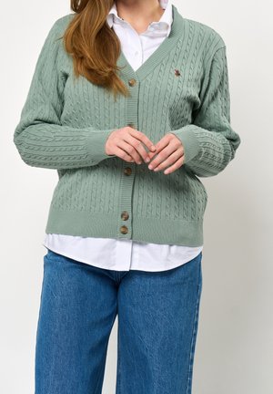 Vrouw met lang bruin haar die een groene kabelgebreide, geknoopte cardigan draagt over een wit overhemd en blauwe spijkerbroek, handen gevouwen voor zich.
