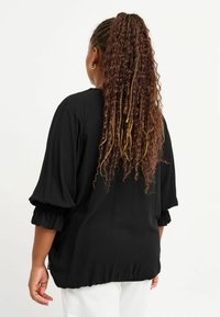 Blusa nera con maniche a sbuffo, polsini arricciati e vestibilità rilassata. Il tessuto ha una consistenza liscia, con un orlo elasticizzato per dare forma.