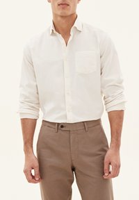 Lys beige skjorte med knapper og en brystlomme, parret med brune bukser. Skjorten har krave og oprulledeærmer.