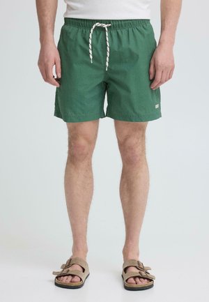 Mężczyzna w zielonych szortach na sznurku i beżowych sandałach stoi na tle gładkiego, jasnego tła.