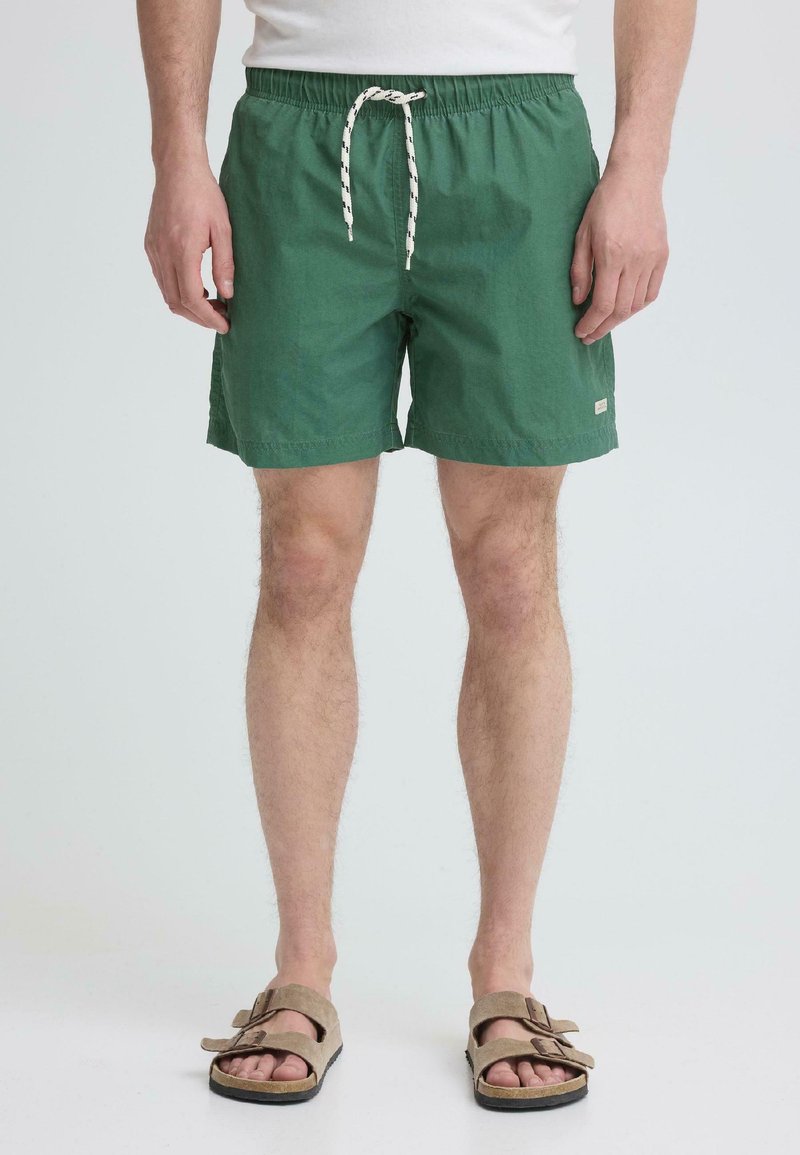 Mees, kes kannab rohelist nööriga lühikesi pükse ja beeže sandaale, seisab ühevärvilise heleda tausta ees.