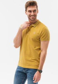 Polo giallo con colletto e dettagli del logo, abbinato a jeans blu. Il tessuto sembra liscio, con maniche corte e una vestibilità rilassata.